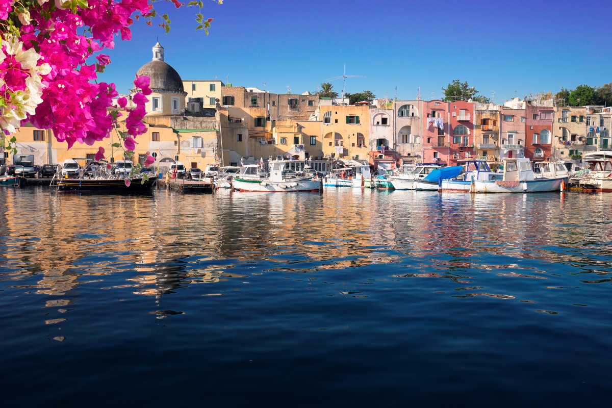 Vista aerea dell'arrivo dei traghetti a Marina Grande, Procida, con case colorate e turisti che sbarcano. Atmosfera accogliente e solare, ideale per trasmettere la facilità dell'arrivo sull'isola.