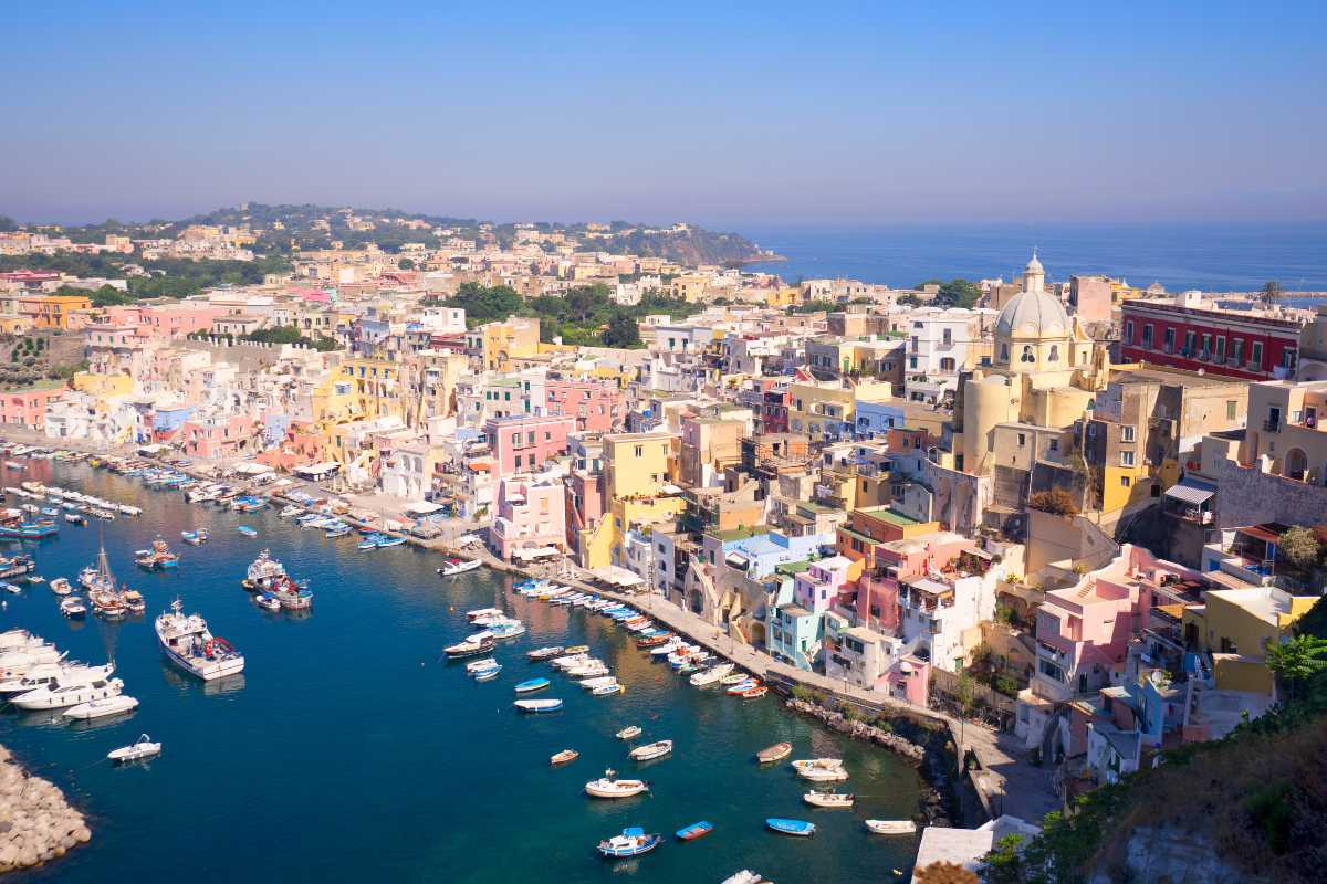 Veduta panoramica dall’alto della Marina di Corricella con le case colorate, barche ormeggiate e il mare turchese; atmosfera estiva e luminosa, Procida