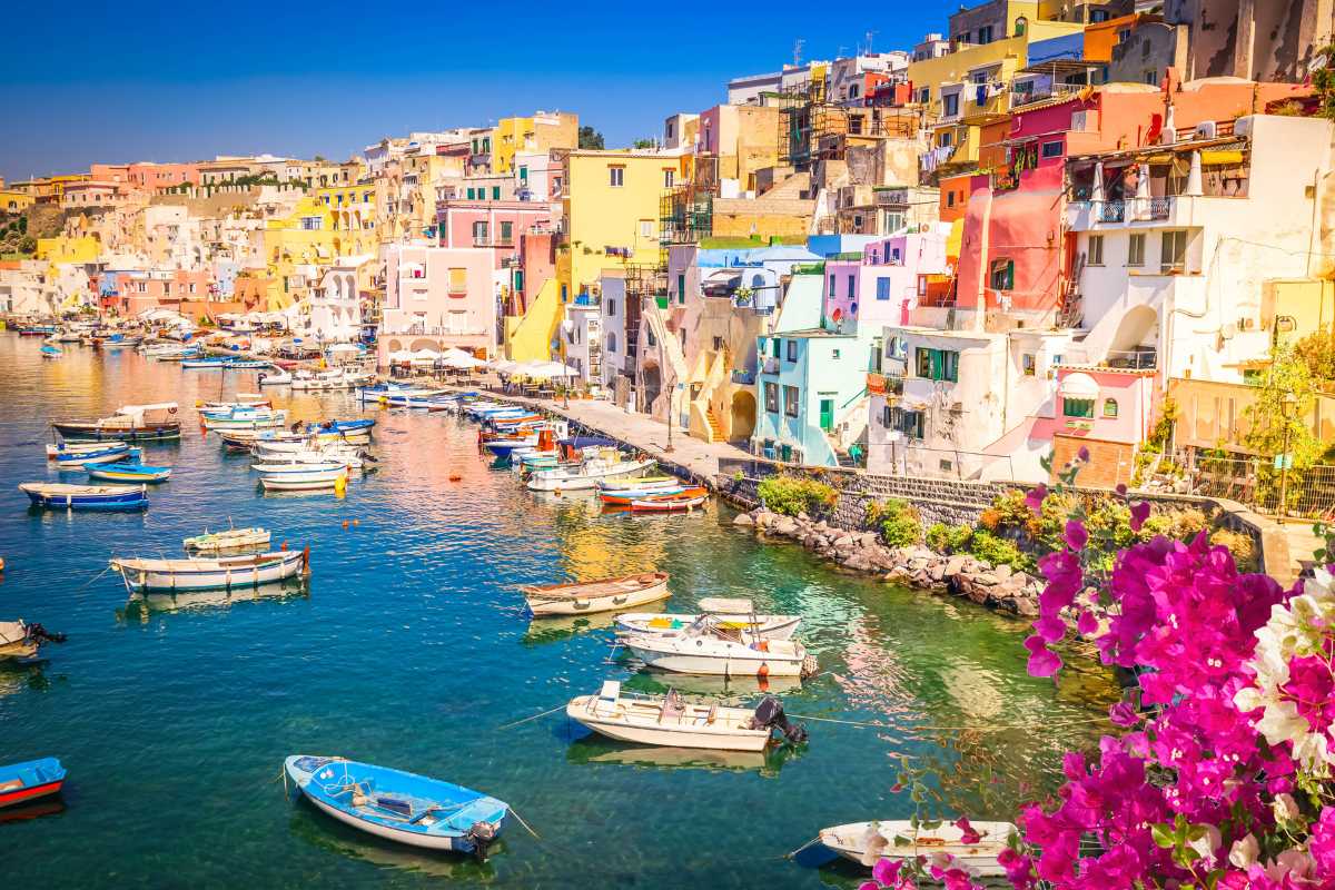 Scorcio di un tavolo apparecchiato con piatti tipici procidani, vista mare sullo sfondo e scorci colorati delle case di Marina Corricella